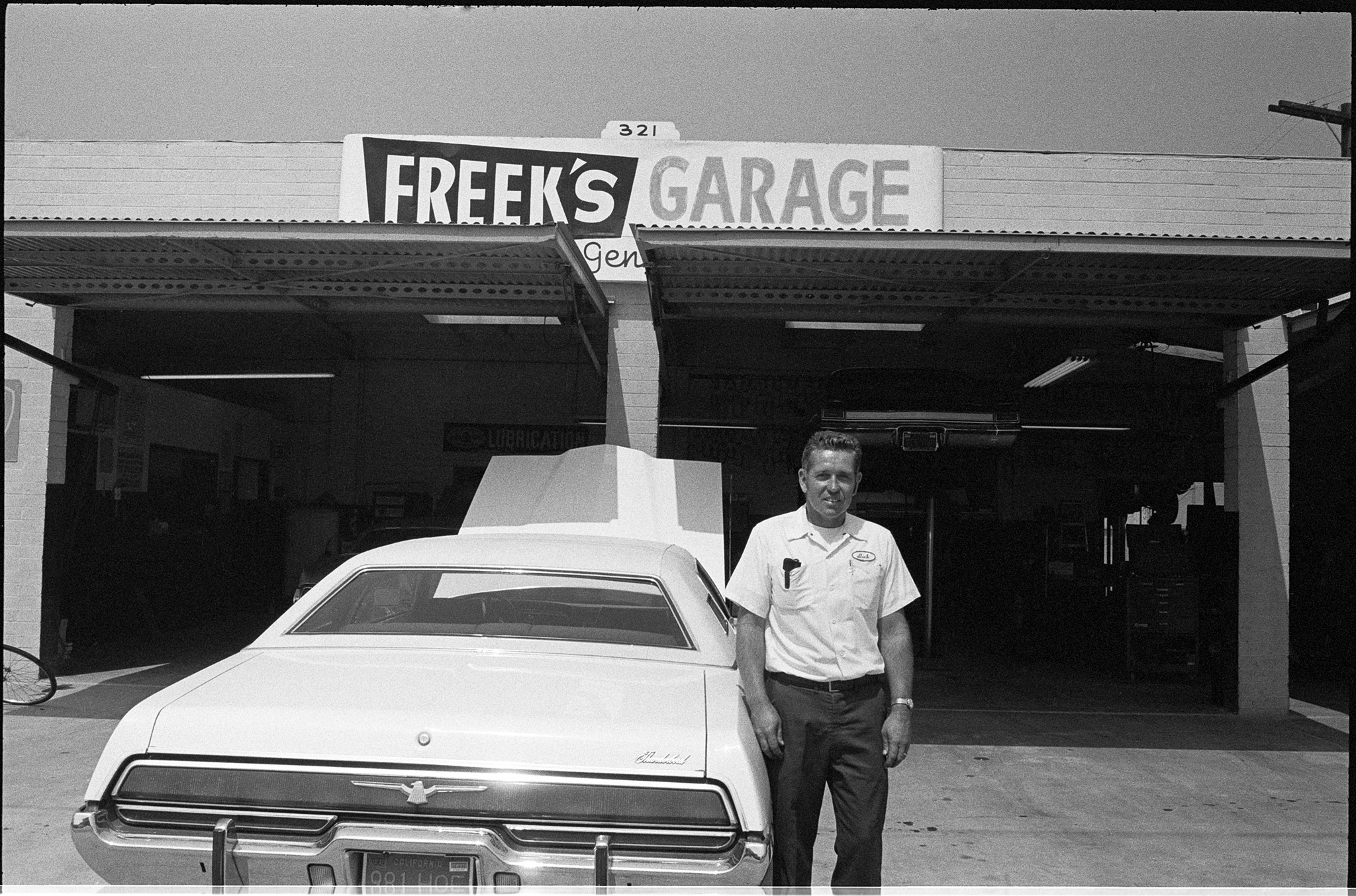 Guy in front of a garage in a white shirt, and black pants. Car with the hood open infront of two