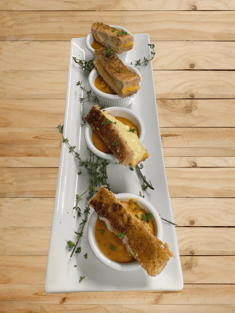 Tiny slices of grilled cheese on top of small bowls of tomato soup on a plate with four selections.