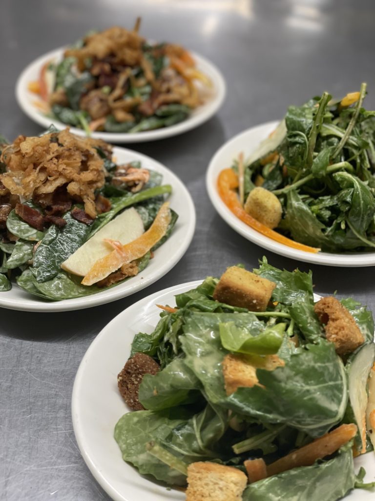 Four small plates of various salads with greens, apples and croutons.