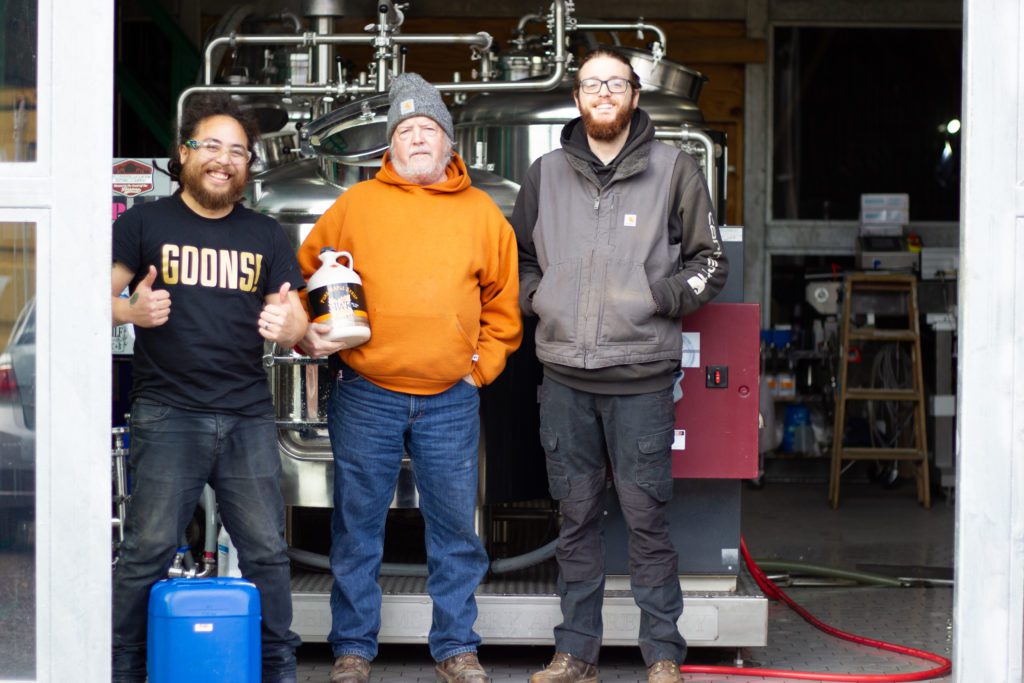 Three working men standing in front of a brewery with the door open to show the brewhouse. One man holds a large bottle of Maple Syrup.