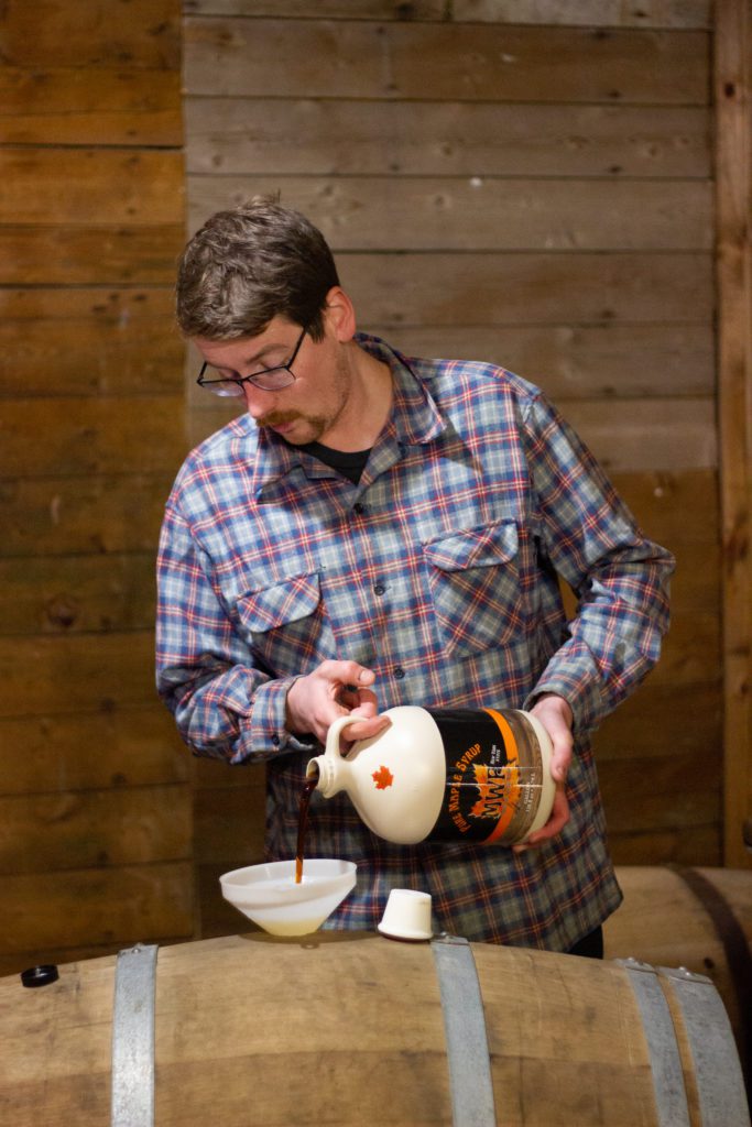 Man stands behind a wooden barren pouring in a large jug of maple syrup