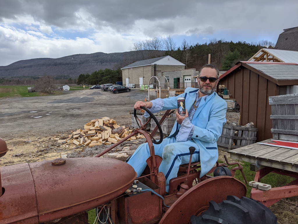 Man in a blue suit sitting on a vintage tractor holding a beer.