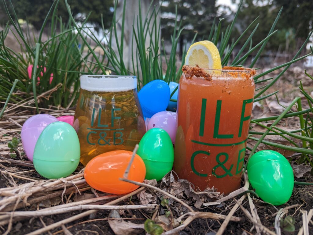 Two ILFC&B glasses, one with beer, and one with a beer cocktail. With multi colored easter eggs around it in the grass.